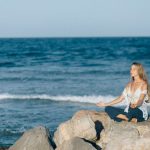 A woman practices meditation outdoors on rocks by the sea, embodying tranquility and relaxation.