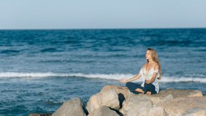 A woman practices meditation outdoors on rocks by the sea, embodying tranquility and relaxation.